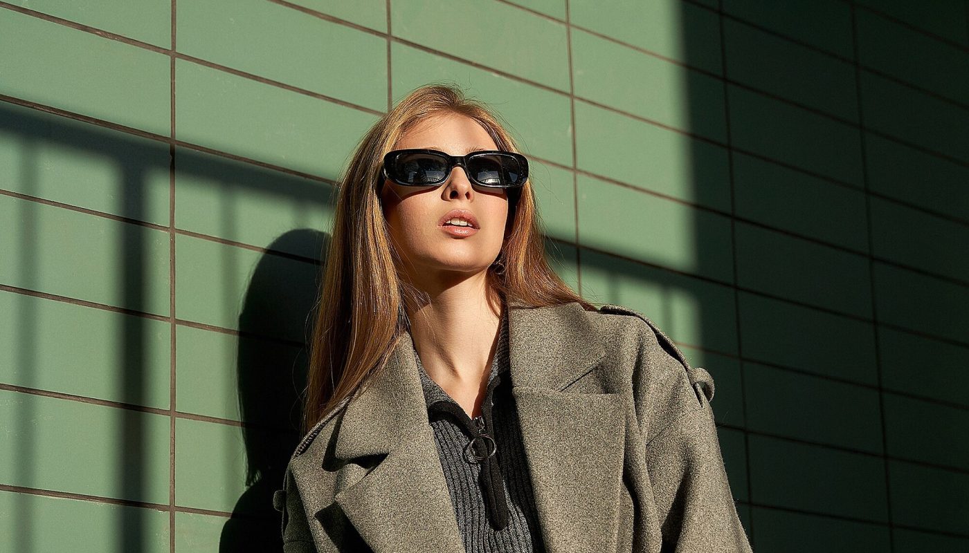 A young woman exudes confidence as she poses in a chic coat, sunglasses on, against a textured green wall. The interplay of shadows creates an artistic ambiance, showcasing high fashion.
