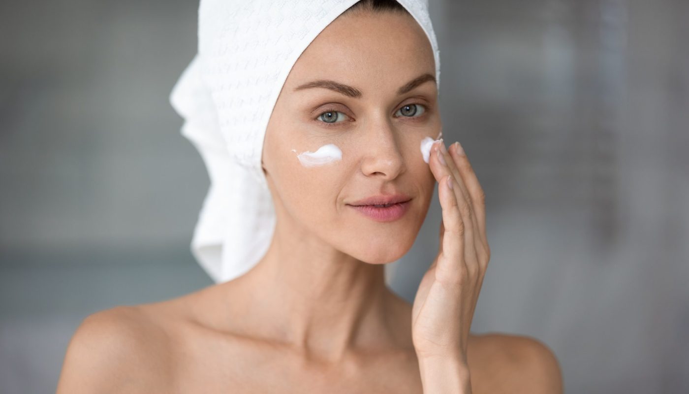 Peaceful attractive young lady with wrapped in towel head applying hydrating moisturizing creme, looking at camera. Beautiful pretty woman doing skincare routine after showering at home or hotel.